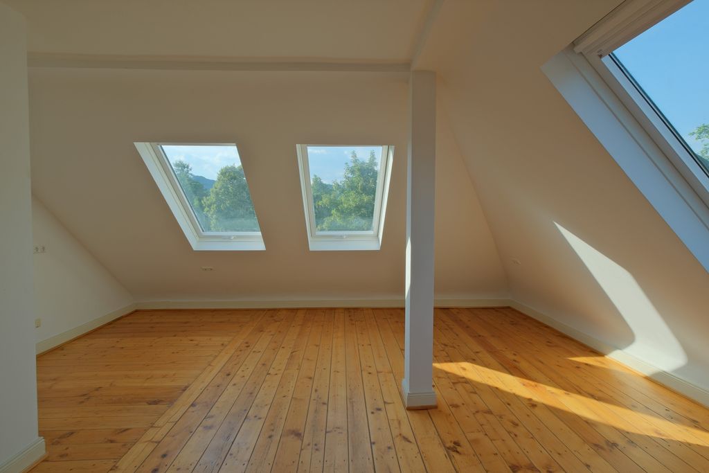 Leeres Dachgeschosszimmer mit großen Dachfenstern, Holzboden und weißen Wänden, durch die Tageslicht einfällt.
