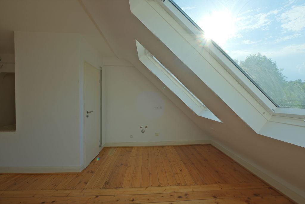 Hell beleuchteter leerer Raum mit schrägem Dachfenster und Holzboden.