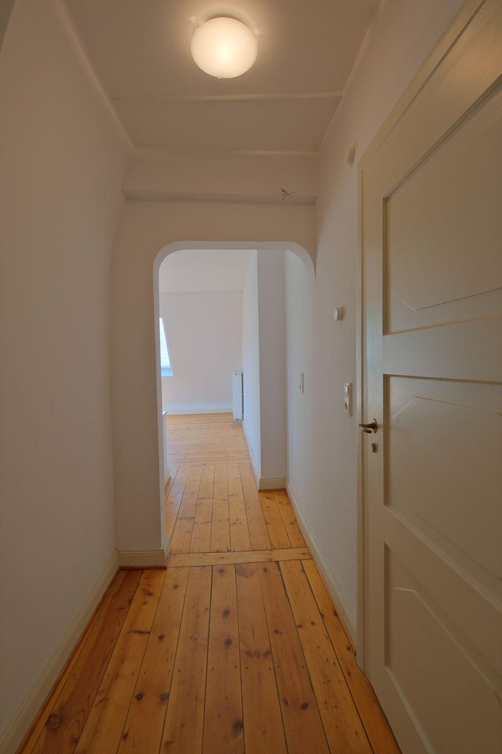 Flur mit Holzboden und weiß gestrichenen Wänden, der durch einen Rundbogen in einen weiteren Raum mit Fenster und Heizung führt.