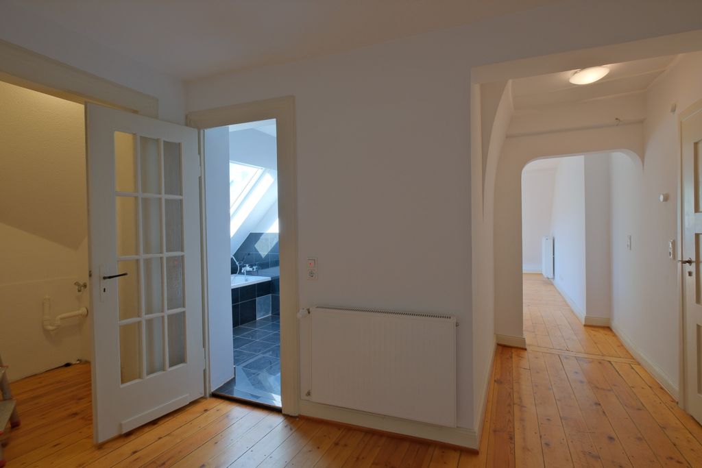 Flur mit heller Holzboden und weißen Wänden, offener Tür zu einem Badezimmer mit schwarzen Kacheln und Badewanne unter einem Dachfenster.