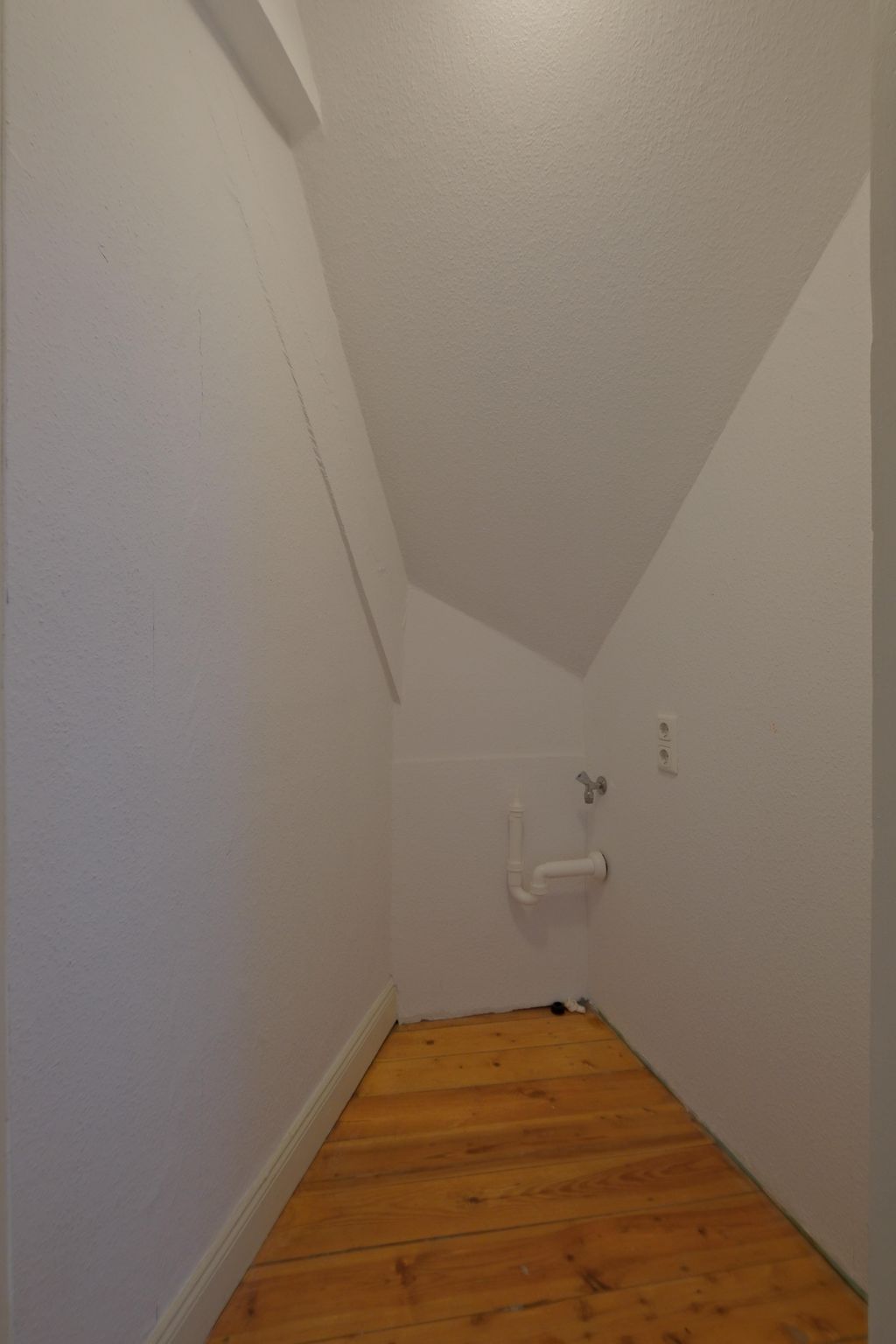 Kleiner leerer Raum mit schräger Decke, weißen Wänden und Holzfußboden, ausgestattet mit einer Wasserleitung und einem Wasserhahn auf der rechten Seite.
