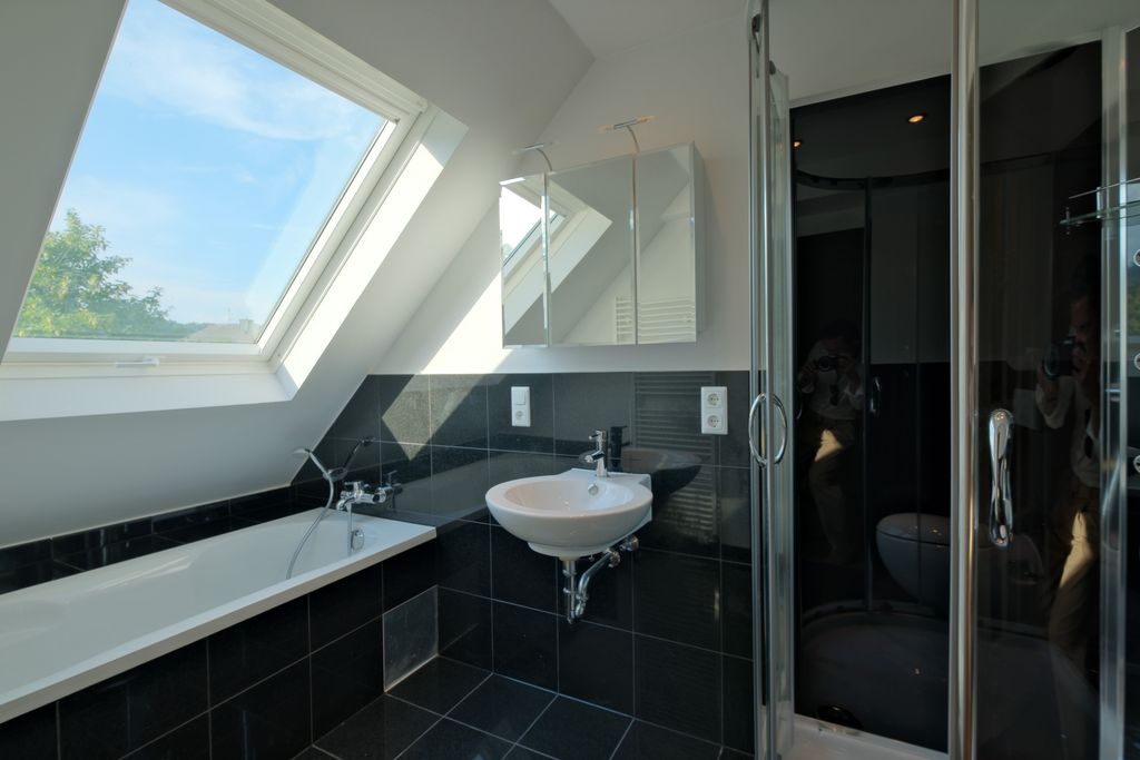 Modernes Badezimmer mit schwarzer Fliesenwand, Badewanne unter einem großen Dachfenster, Waschbecken und Glastür zur Dusche.