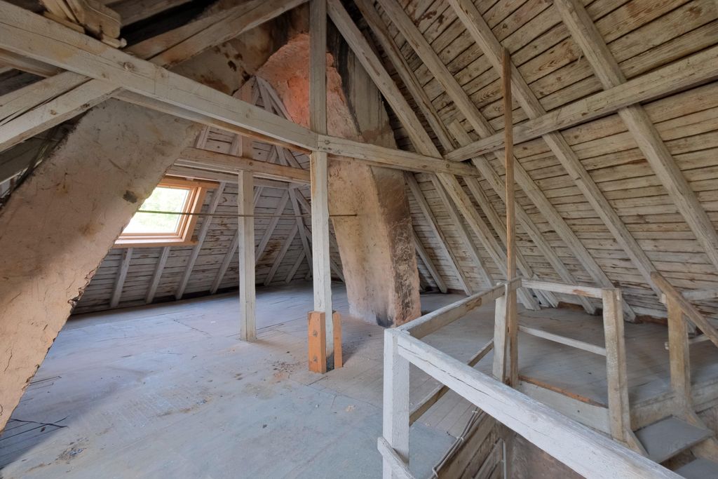 Dachboden mit sichtbaren Holzbalken, einer Dachluke und einem großen verputzten Schornstein im Raum.