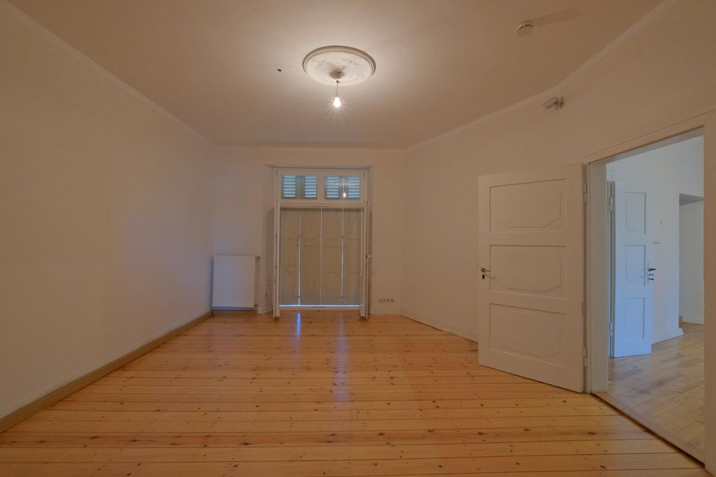 Leerer Raum mit Holzboden, weißen Wänden, einer offenen weißen Tür rechts und einem Fenster mit geschlossenen Lamellen am Ende.
