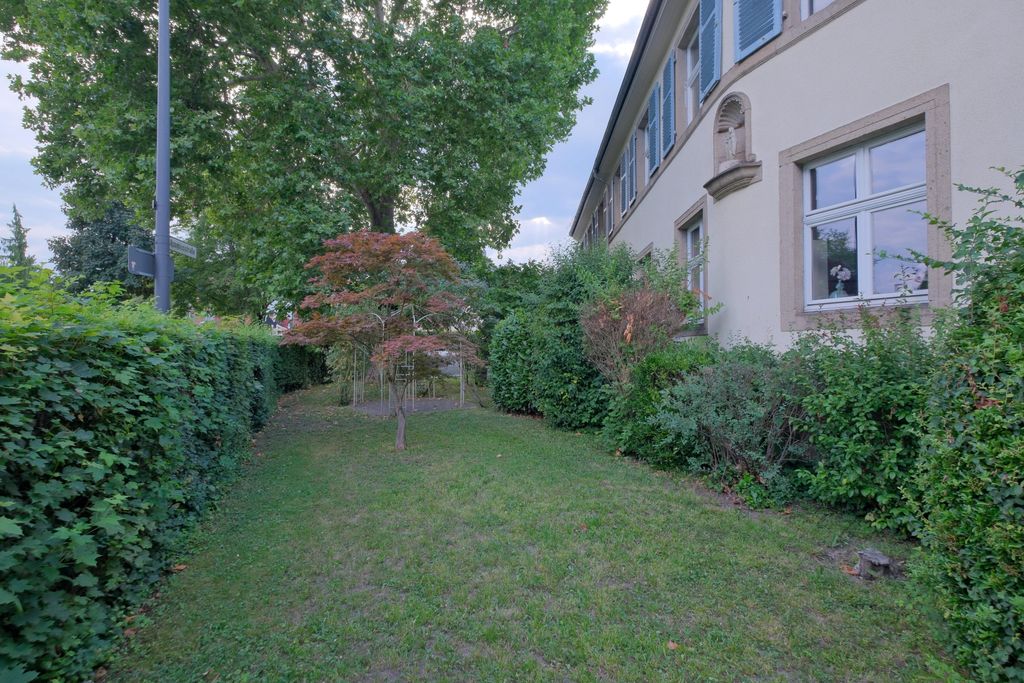 Grüner Gartenbereich neben einem hellen Gebäude mit blauen Fensterläden, umgeben von Büschen und Bäumen.