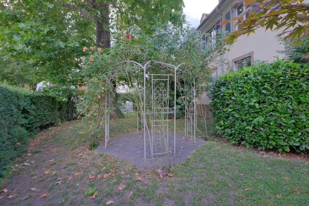 Ein leerer Pavillon aus weißem Metall mit überwachsenen Kletterpflanzen in einem Garten neben einem Gebäude.