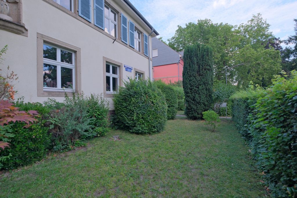 Ein gepflegtes grünes Gartengrundstück neben einem hellen Gebäude mit blauen Fensterläden, umgeben von verschiedenen Sträuchern und Bäumen unter blauem Himmel.
