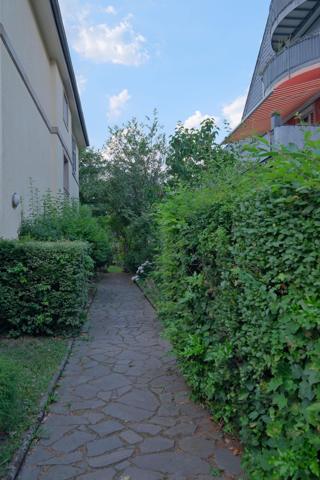 Ein gepflasterter Gehweg zwischen einem Wohngebäude und einer hohen, grünen Hecke unter einem blauen Himmel mit einigen Wolken.