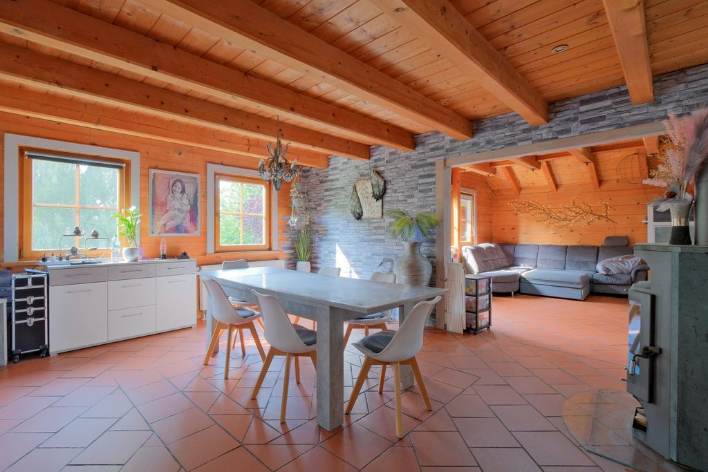 Moderne Wohnküche mit Holzbalkendecke, großem Esstisch, weißen Stühlen und einem angrenzenden Wohnzimmer mit grauem Ecksofa in einem Raum mit Holz- und Steinwänden.