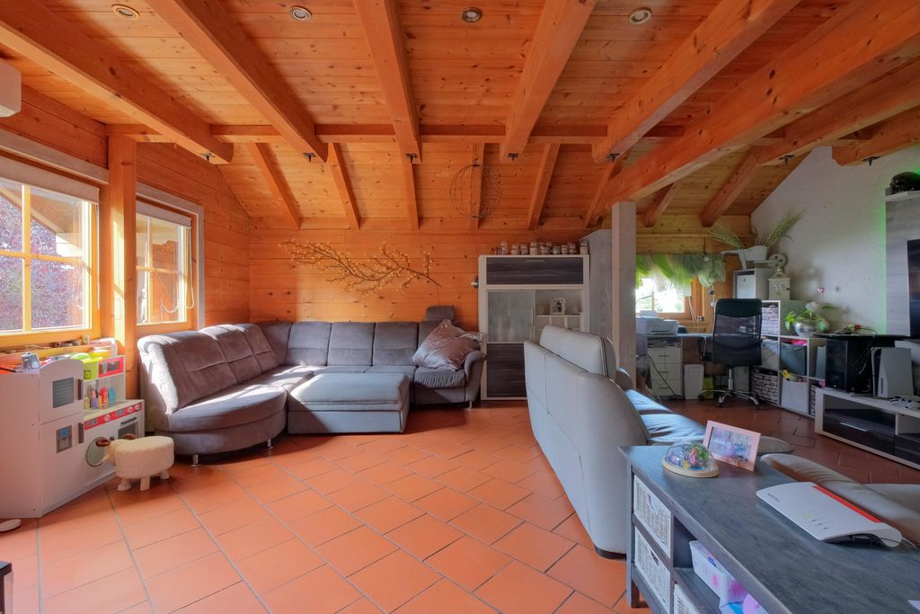 Großes Wohnzimmer mit Holzwänden und sichtbaren Dachbalken, grauem Ecksofa und an der Wand dekorierter Zweig.