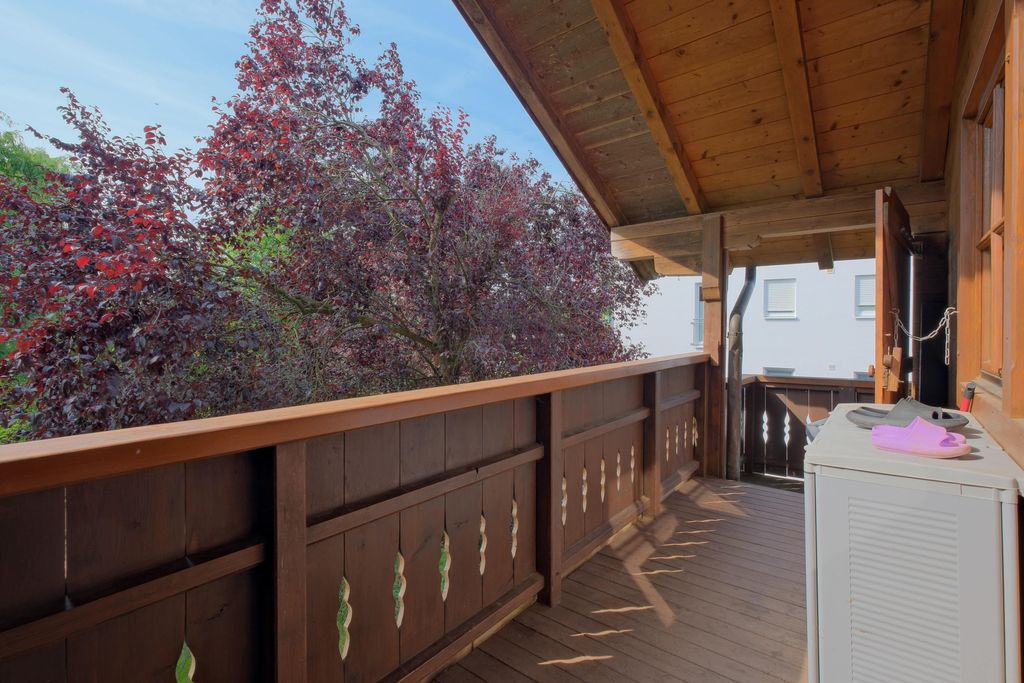 Balkon mit braunem Holzgeländer und einem überdachten Dach, daneben ein rotes Laub-Baum und ein weißes Gebäude im Hintergrund.