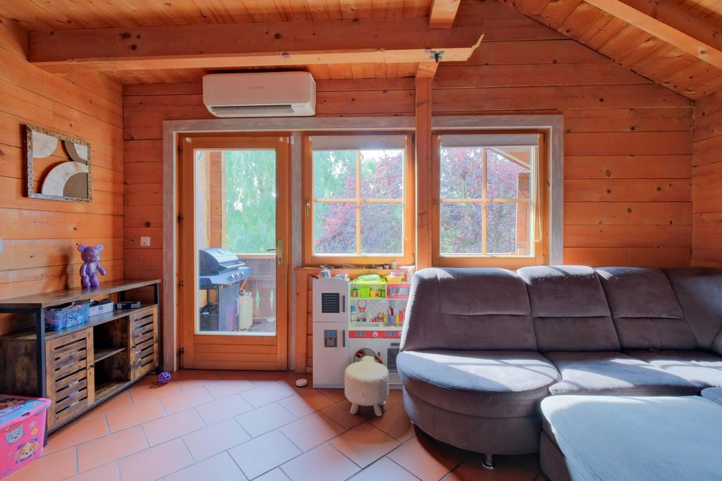 Innenraum eines gemütlichen Holzhauses mit großem Fenster, grauem Stoffsofa, Spielküche für Kinder und Terrasse mit Grill im Hintergrund.