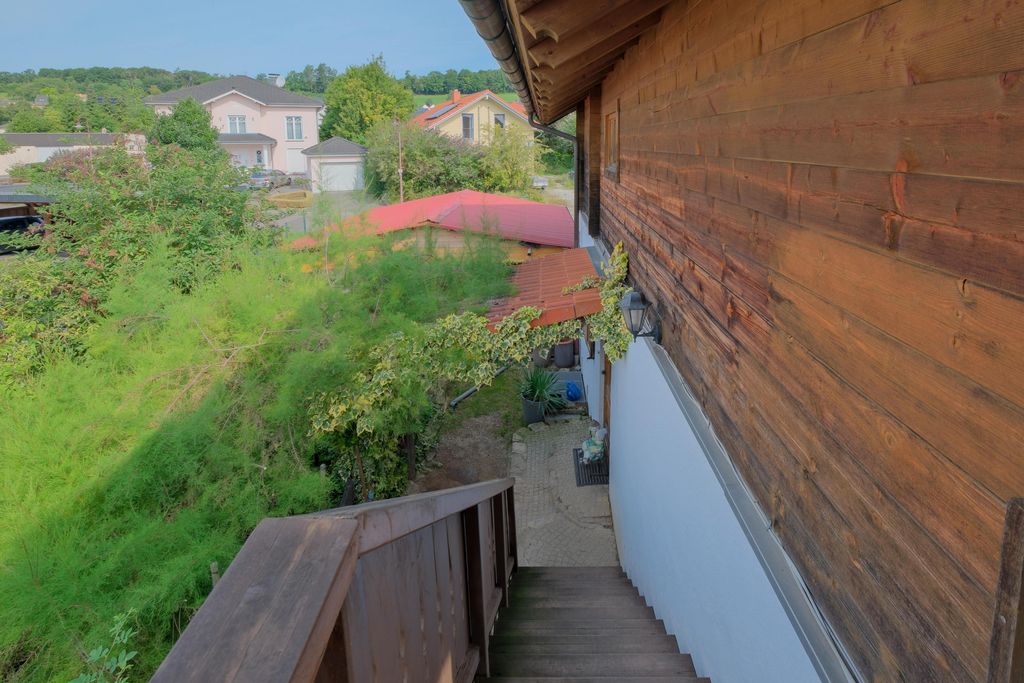 Holztreppe neben einem Haus mit Holzverkleidung, umgeben von grünen Büschen und Blick auf weitere Häuser im Hintergrund.