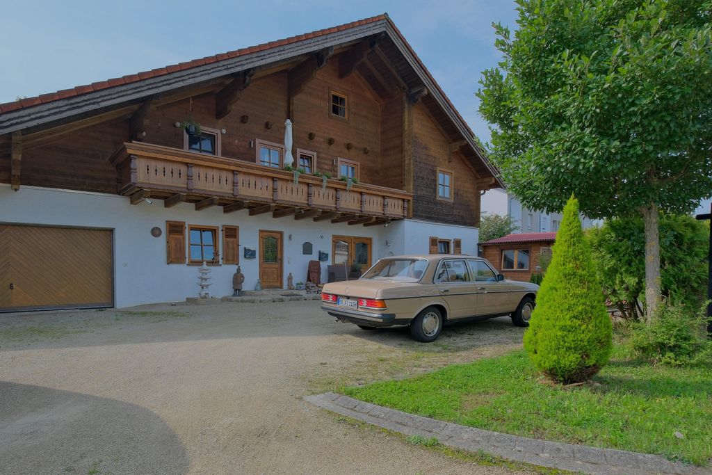 Ein klassisches älteres Auto parkt vor einem traditionellen, zweistöckigen Haus mit Holzverkleidung und einem Balkon, umgeben von einem kleinen Garten mit Bäumen.