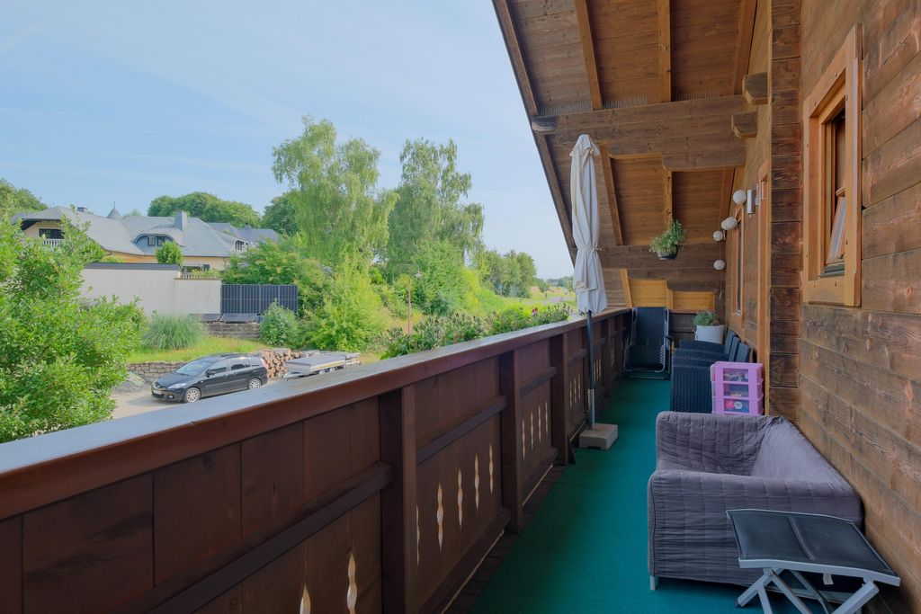 Blick von einem langen Balkon mit Holzboden und Holzgeländer auf grün bewachsene Häuser und eine Straße mit parkendem Auto an einem sonnigen Tag.