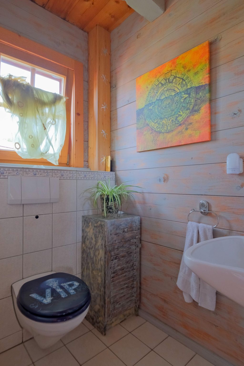 Kleines Badezimmer mit Holzwänden, hängender Toilette mit einem Toilettensitz mit der Aufschrift "VIP", einem kleinen Schrank mit einer Pflanze und einem Handtuch neben einem Waschbecken.