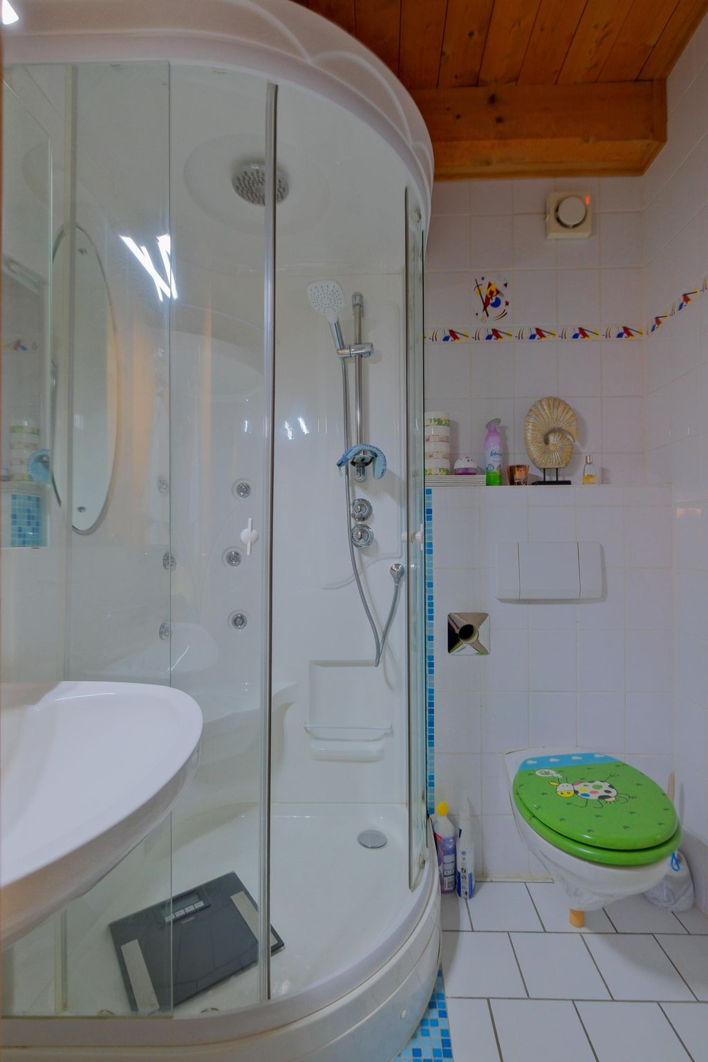 Kleines Badezimmer mit einer runden Duschkabine, einem weißen Waschbecken und einer Toilette mit grünem Deckel und einem bunten Marienkäfer-Motiv, weiße Fliesenwände mit einer bunten Bordüre und eine Holzbalkendecke.
