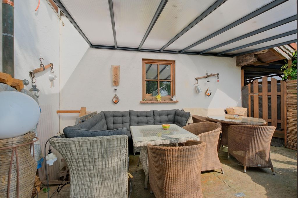 Überdachte Terrasse mit graßem grauem Ecksofa, einem Glas-Couchtisch sowie mehreren Korbsesseln und einem runden Tisch vor einer weiße Wand mit Fenster.