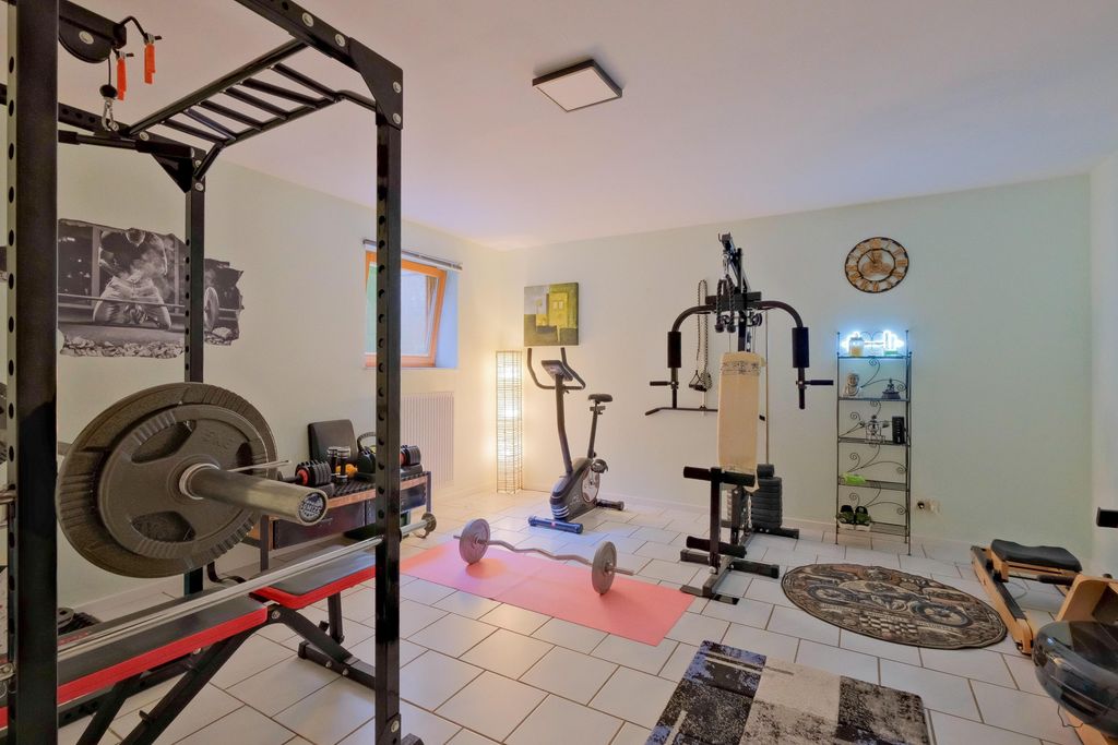 Ein gut ausgestatteter Fitnessraum in einem hellen Raum mit verschiedenen Trainingsgeräten wie einem Hantelständer, Fitnessbike, Gewichtszugmaschine und einer Hantelstange auf einer Yogamatte.