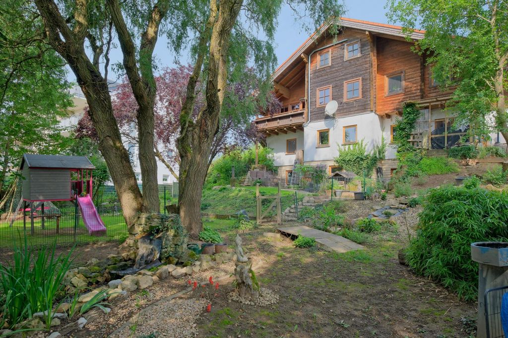 Großes Holz- und Steinhaus mit einem Garten, Rutsche und Spielplatz im Vordergrund, umgeben von Bäumen und Pflanzen.