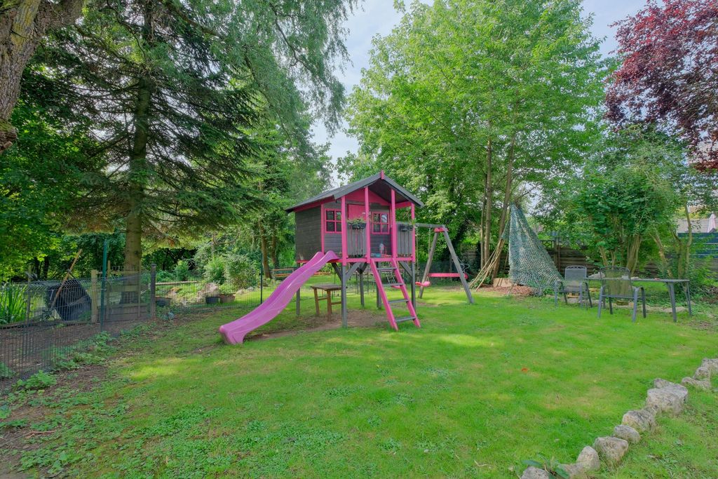Kinderspielplatz im Garten mit einem hölzernen Spielhaus, einer rosa Rutsche, Schaukel und Kletternetz, umgeben von Bäumen und Grünflächen.