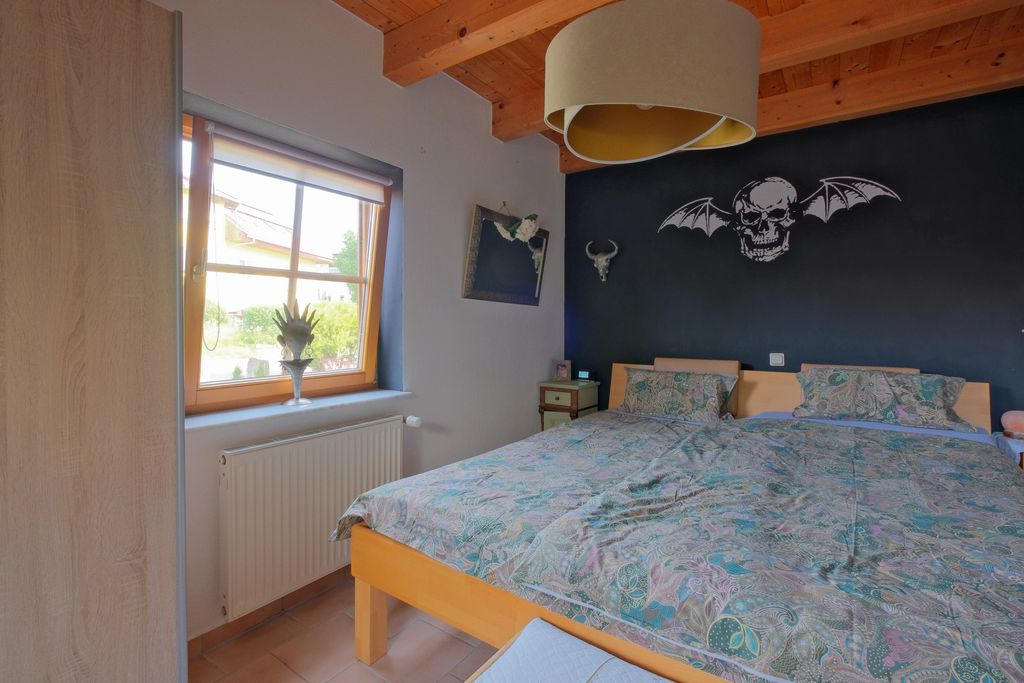 Schlafzimmer mit Doppelbett und floral gemusterten Bettbezügen, schwarzer Wand mit Totenkopf mit Fledermausflügeln als Wanddekoration und Fenster mit Blick nach draußen.