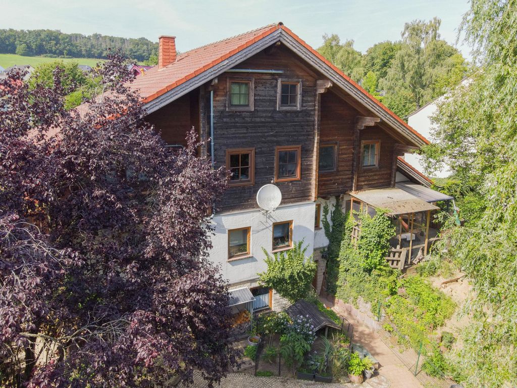 Ein traditionelles mehrstöckiges Holzhaus mit rotem Ziegeldach, umgeben von grünen Bäumen und Pflanzen in einer ländlichen Umgebung.