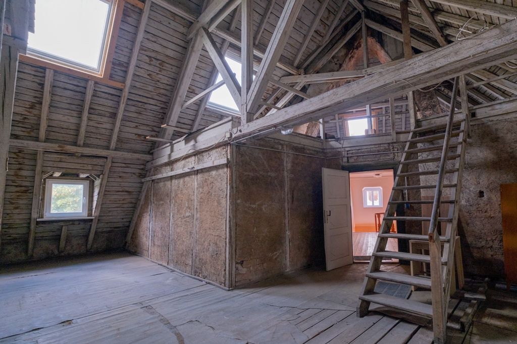 Alter, leerer Dachboden mit sichtbaren Holzbalken, einer kleinen Treppe und offenem Tür zur hellen, gegenüberliegenden Wand mit Fenster.