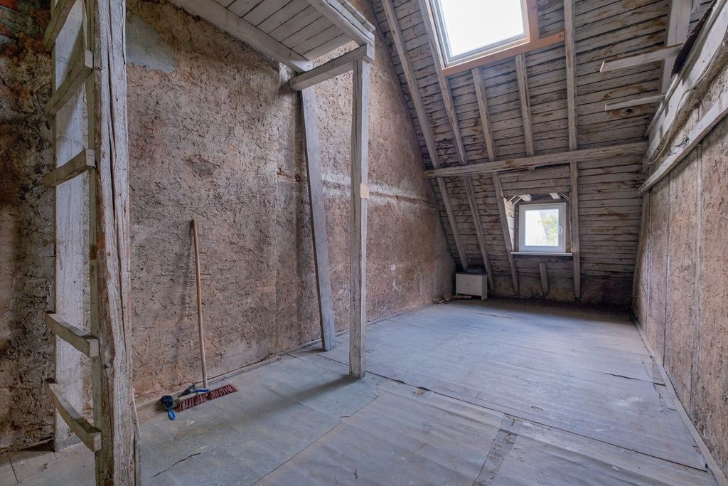Ein leerer, alter Dachboden mit schrägen Holzwänden und einem Fenster im Giebeldach, mit sichtbaren Holzbalken und einem Besen, der an der Wand lehnt.