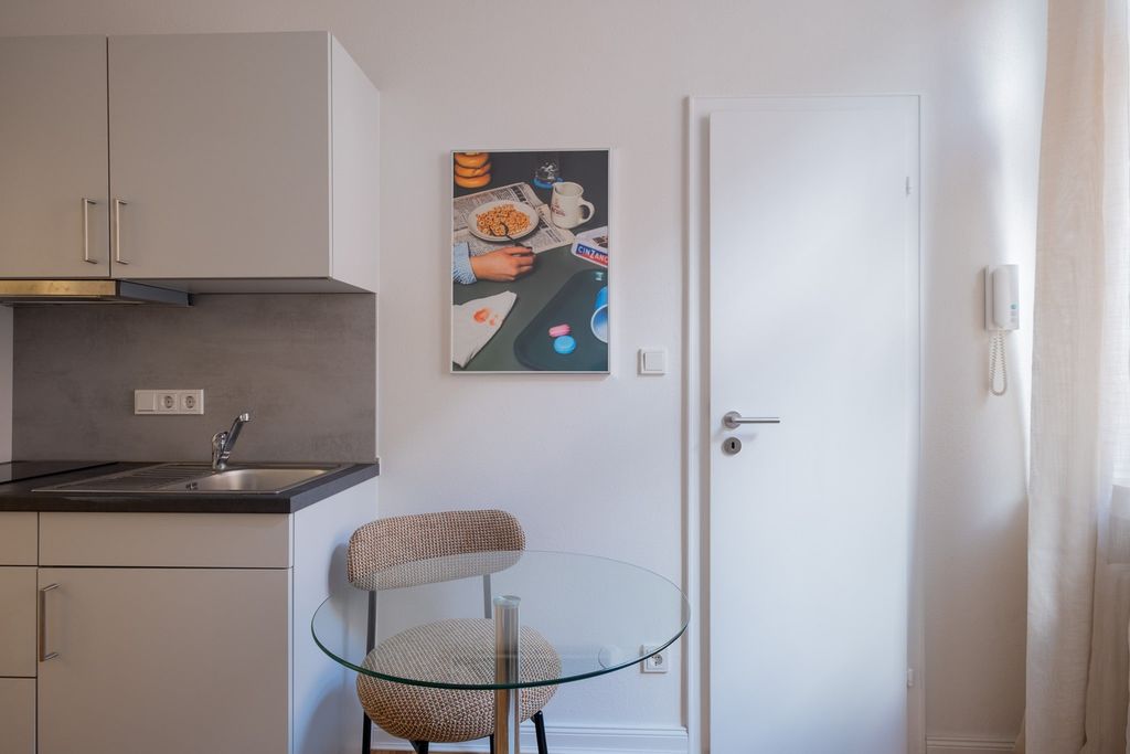 Kleine, moderne Küchenzeile mit weißen Schränken und Spüle, daneben ein runder Glastisch mit gepolstertem Stuhl vor einer weißen Tür und einem Bild an der Wand.