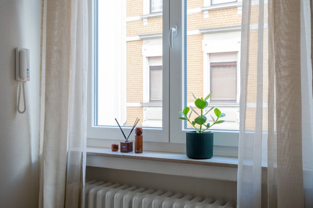 Ein Fenster mit halbtransparenten Vorhängen, einem Pflanzenkübel und Aromastäbchen auf der Fensterbank, mit Blick auf ein gegenüberliegendes Gebäude.
