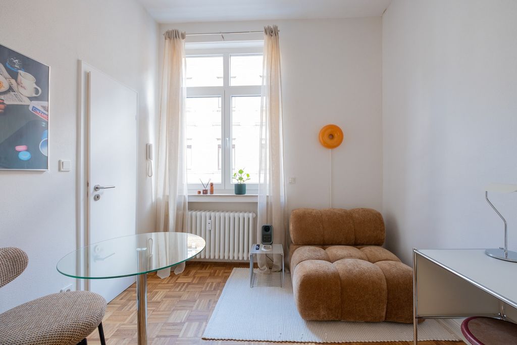 Kleiner, heller Wohnraum mit braunem, gepolstertem Sessel, rundem Glastisch und großer Fensterfront mit weißen Vorhängen.