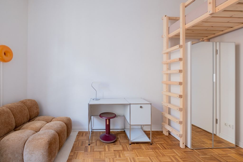 Minimalistisch eingerichteter Raum mit einem braunen Sofa, einem weißen Schreibtisch mit rotem Hocker und einer hölzernen Hochbettleiter neben einem großen Spiegel.