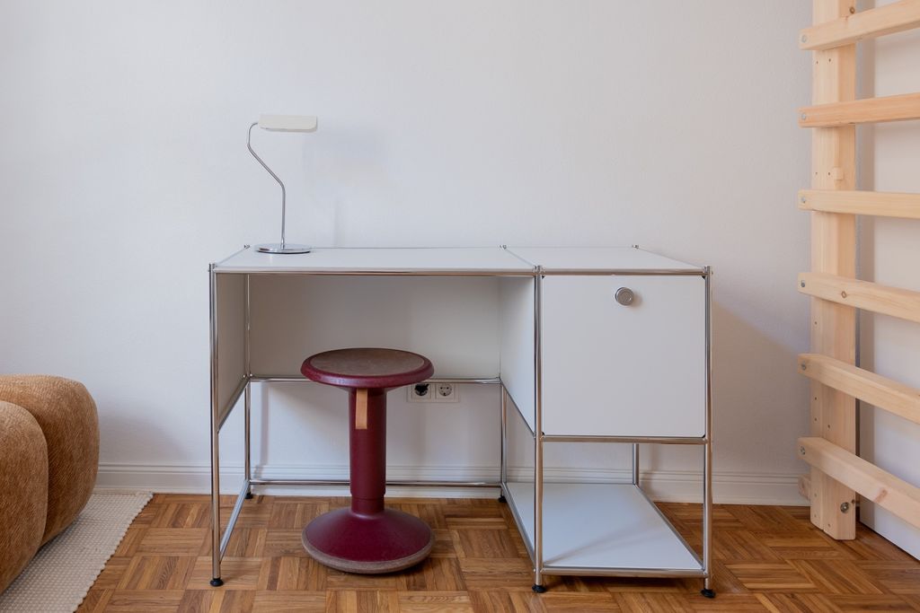 Ein moderner, weißer Schreibtisch mit einem roten Stehhocker in einem Raum mit Parkettboden, einer beige Couch links und einer Holzleiter rechts.