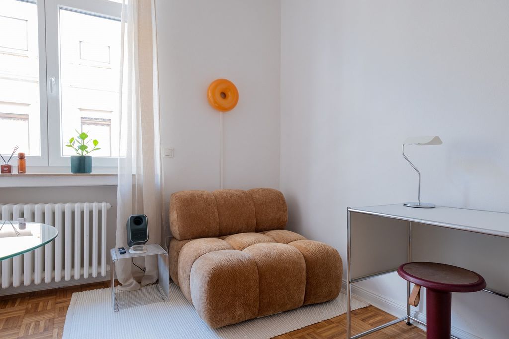 Helles Wohnzimmer mit einem braunen, gepolsterten Sessel neben einem weißen Schreibtisch und einem kleinen runden Hocker. An der Wand hängt eine orangefarbene Wandlampe, gegenüber einem Fenster mit durchsichtigen Vorhängen.