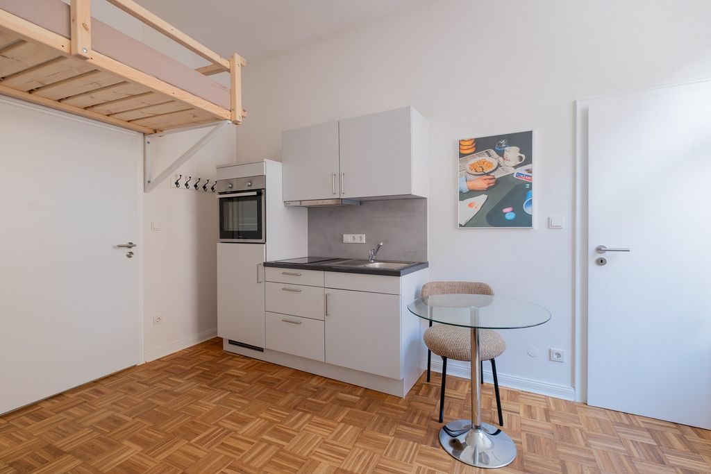 Kleine moderne Küchenzeile in einem Raum mit weißen Wänden, Parkettboden und einem runden Glastisch mit beige gepolstertem Stuhl. Über der Küchenzeile hängt ein Bild mit einer Illustration von Frühstücksszene.
