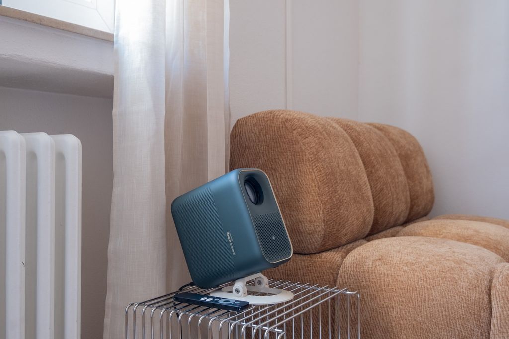 Kleiner blauer Projektor auf einem kleinen Metallgittertisch neben einem braunen, gepolsterten Sofa in einem hellen Raum mit Gardinen und Heizkörper.