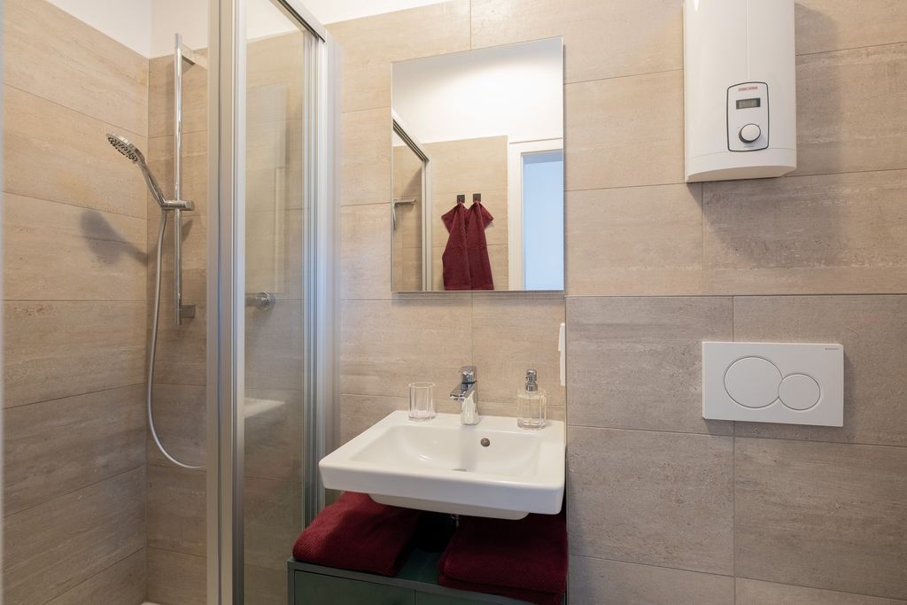 Ein modernes Badezimmer mit einer Eckdusche, einem weißen Waschbecken mit Spiegel darüber, zwei dunkelroten Handtüchern unter dem Waschbecken und einem weißen Durchlauferhitzer an der Wand.
