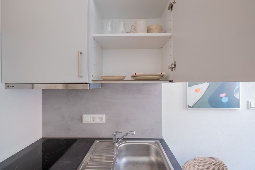 Offener Küchenschrank über einer Spüle mit kleinen Tellern, Tassen und Gläsern, in einer modernen Küche mit grauer Rückwand und schwarzer Arbeitsplatte.