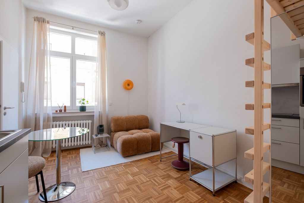 Kleines Wohnzimmer mit einem braunen Sesselsessel, einem runden Glastisch, einem weißen Teppich und einem weißen Schreibtisch in einem Apartment mit Holzboden und großen Fenstern.