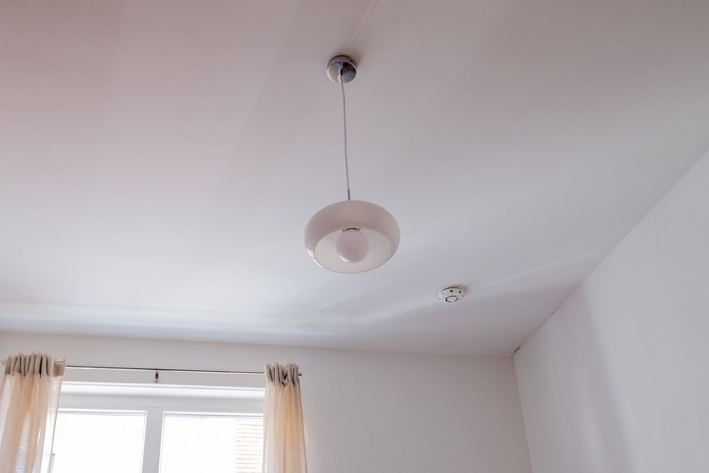 Blick an die weiße Decke eines Raums mit einer hängenden weißen Deckenlampe, einem Rauchmelder und einem Fenster mit halbtransparenten Vorhängen.