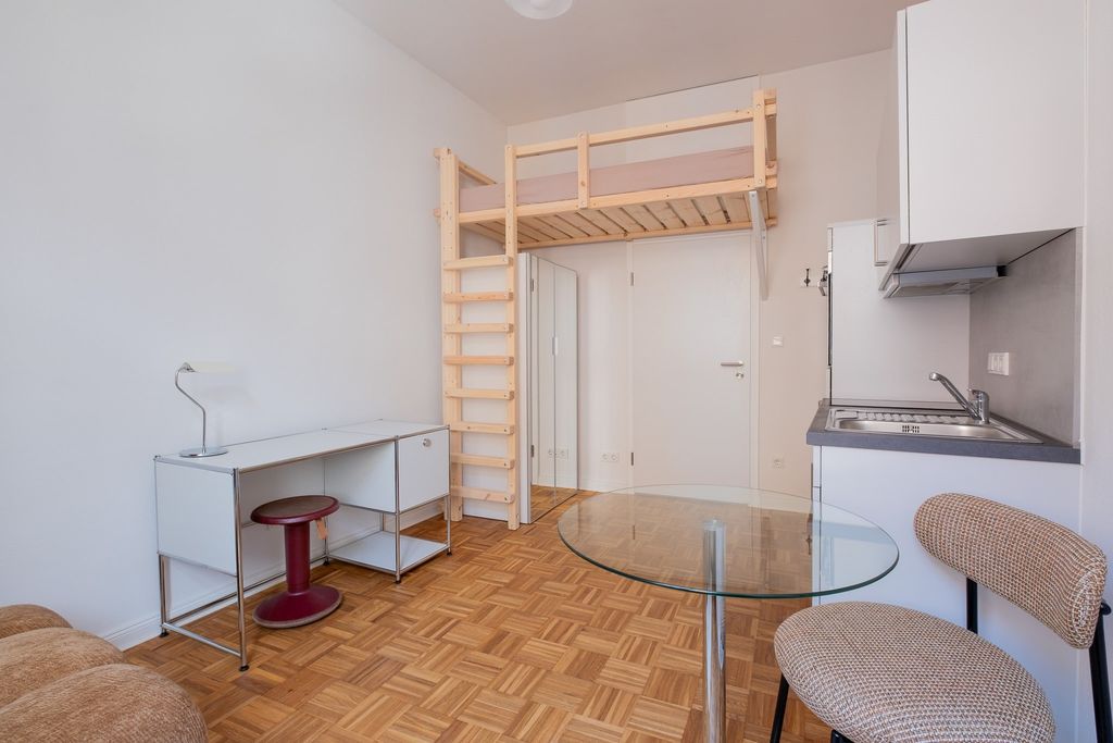 Kleines Zimmer mit Hochbett aus Holz, weißem Schreibtisch, rotem Hocker, Glas-Tisch und beige gepolstertem Stuhl, angrenzender Küchenzeile und Parkettboden.
