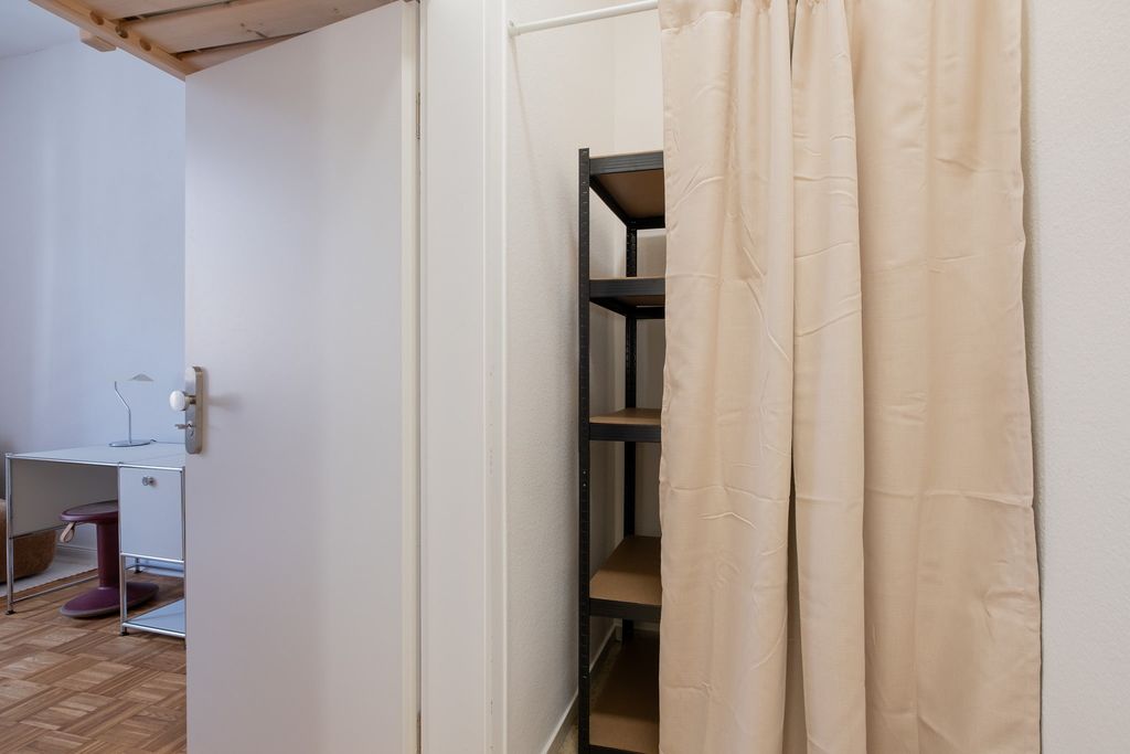 Ein offenes weißes Türblatt mit Blick auf einen Schreibtisch mit einer Lampe und einem roten Hocker in einem Raum, daneben ein Regal mit vier Brettern, verdeckt durch einen beigen Vorhang.