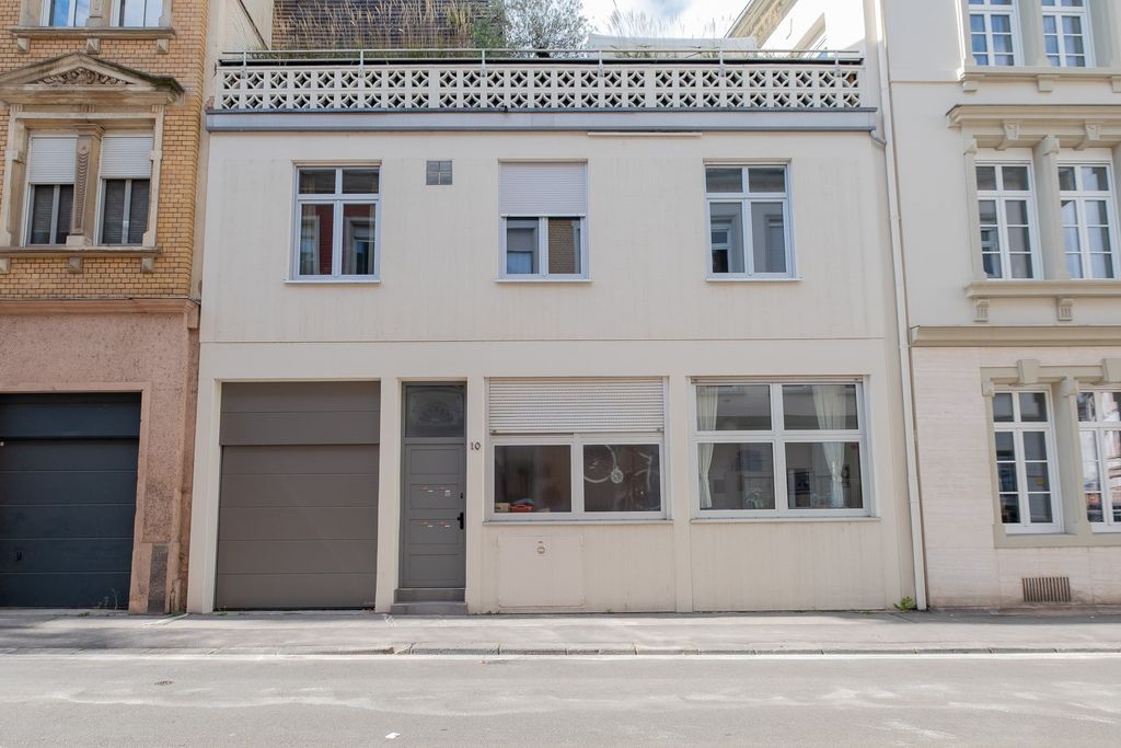 Eine weiße zweistöckige Hausfassade mit mehreren Fenstern, darunter auch Rollläden, und einer grauen Tür mit der Hausnummer 10. Rechts und links sind andere Gebäude sichtbar, und vor dem Haus verläuft eine leere Straße.