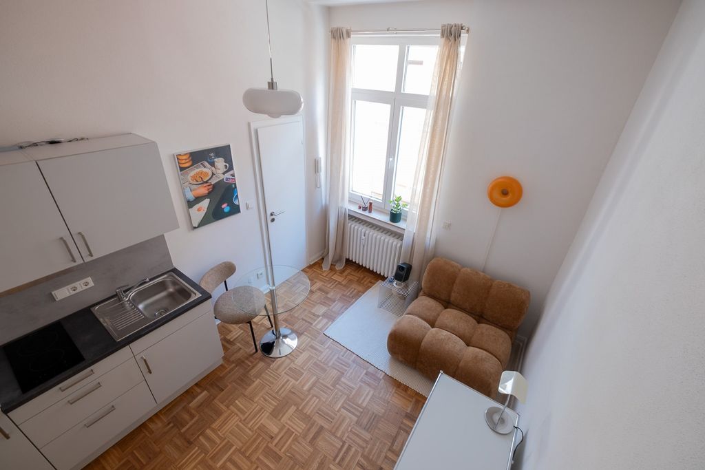Kleines, helles Apartment mit Küchenzeile, kleinem Esstisch mit zwei Stühlen, einem gemütlichen braunen Sessel, Fenster mit Vorhängen und Holzboden.