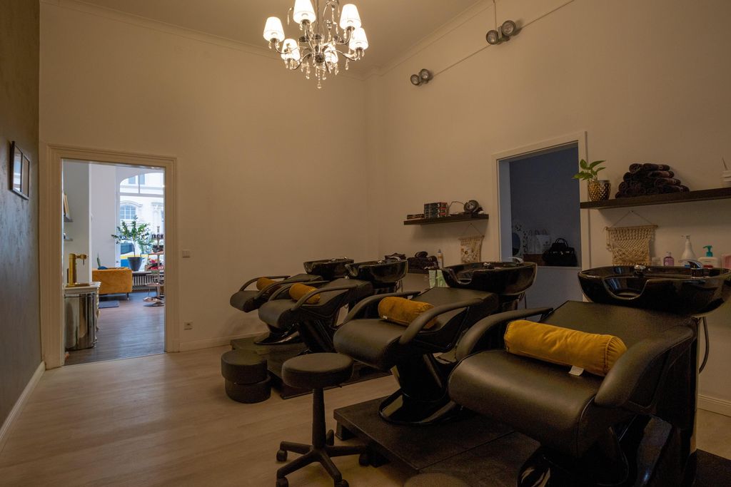 Innenansicht eines Friseursalons mit mehreren schwarzen Waschstühlen, die mit gelben Kissen ausgestattet sind, und einem Kronleuchter an der Decke.