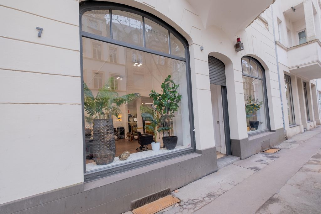 Straßenseite mit großen bogenförmigen Fensterfronten eines Gebäudes, in denen Zimmerpflanzen und Inneneinrichtung sichtbar sind.