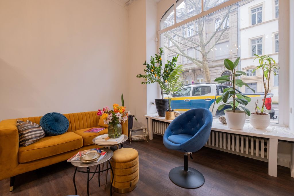 Gemütliches Wohnzimmer mit einem orangefarbenen Samtsofa, einem blauen Samtstuhl und mehreren Pflanzen auf der Fensterbank eines großen Fensters mit Blick auf eine Straße.