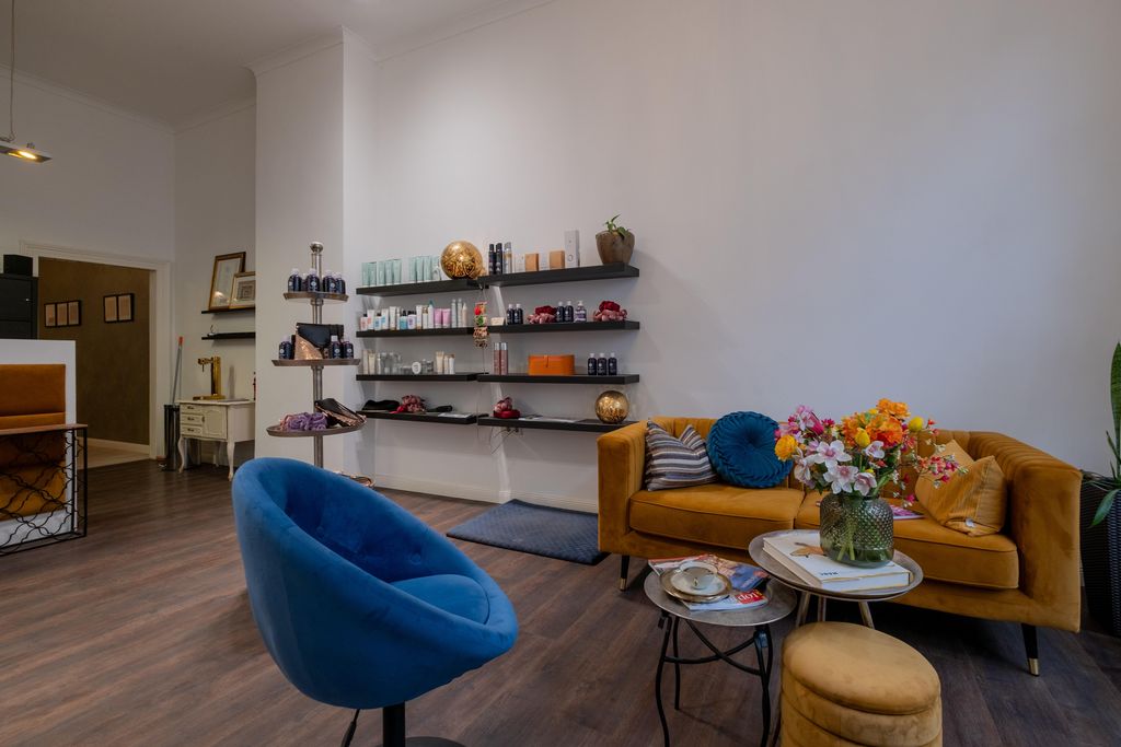 Ein gemütlich eingerichteter Wartebereich mit einem blauen Sessel, einem orangefarbenen Sofa, einem kleinen Couchtisch mit Zeitschriften und einer Vase mit bunten Blumen. An der Wand sind Regale mit verschiedenen Kosmetik- und Pflegeprodukten angebracht.