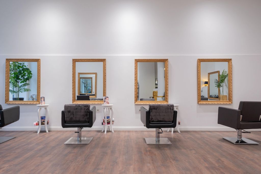 Innenraum eines modernen Friseursalons mit vier Friseurstühlen vor großen Spiegeln an der Wand, kleinen weißen Tischen dazwischen und Holzboden.